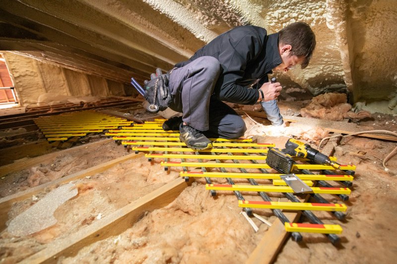 Auditor kneeling in an attic with a flashlight, gloves, and tools, inspecting insulation between exposed beams