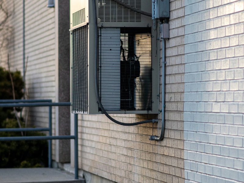 Outdoor HVAC condenser unit mounted on a brick wall with electrical disconnect box and refrigerant pipes