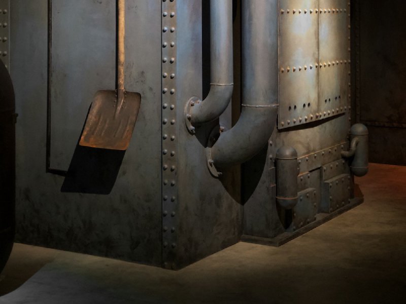 Industrial gas boiler with riveted metal panels, vertical pipes, and gauge in a basement utility room
