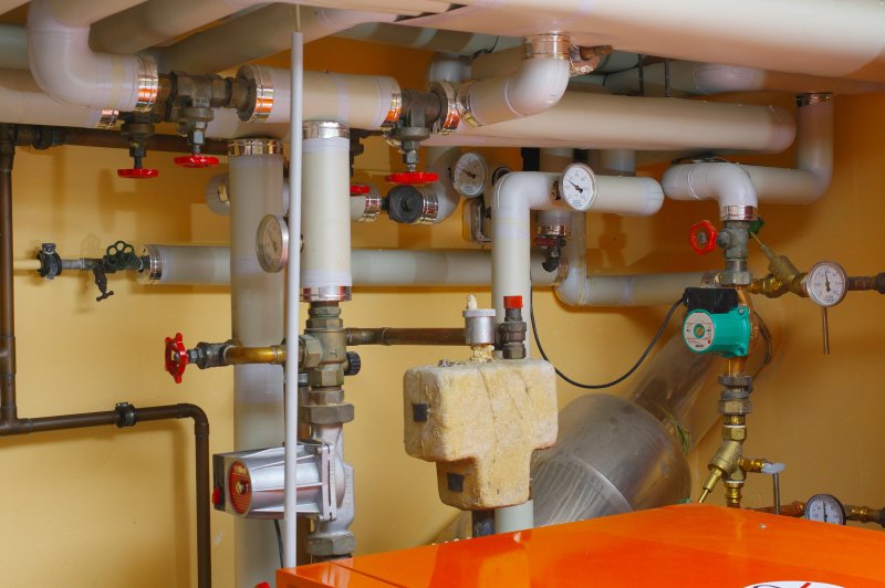 Residential heating system mechanical room with PVC pipes, brass valves, and pressure gauges mounted on a yellow wall