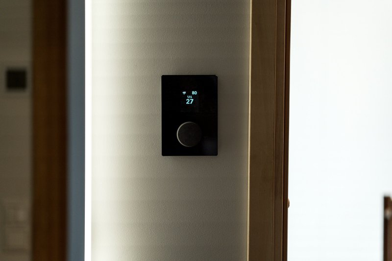 Black smart thermostat mounted on a white wall displaying current temperature and heating mode