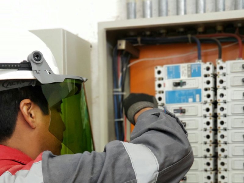HVAC technician in hard hat and overalls testing an open electrical panel with a yellow multimeter