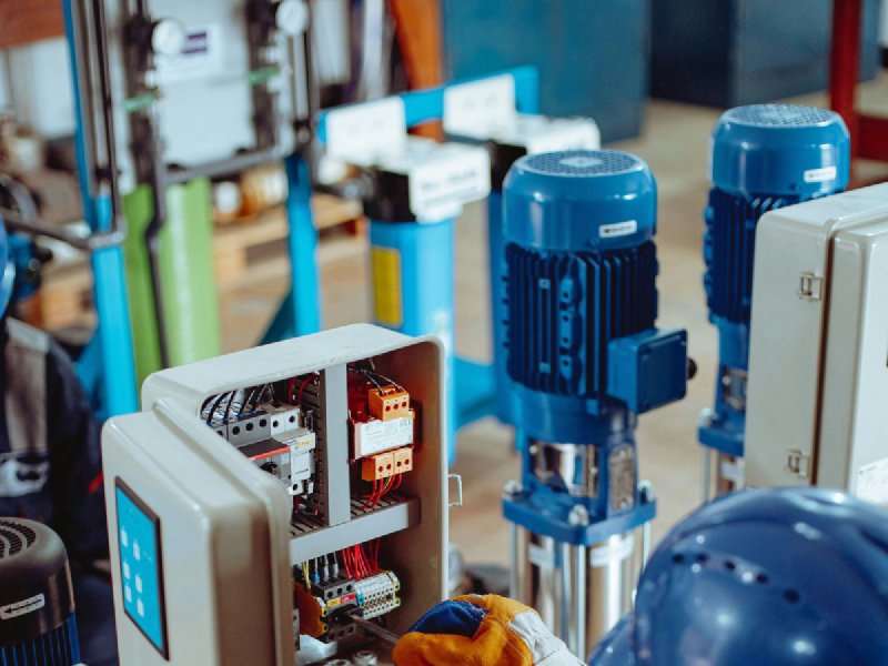 Technician in hard hat and gloves repairing an open electrical control panel with internal wiring exposed