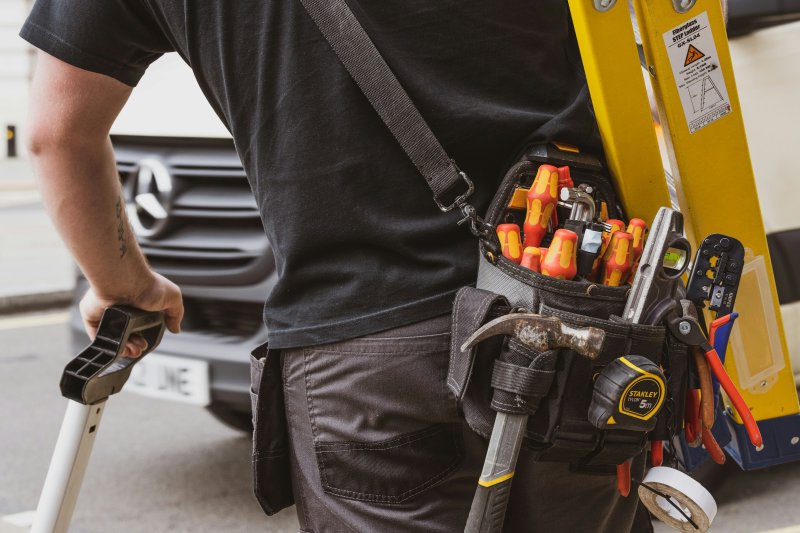 Service technician viewed from behind with a loaded tool belt and a step ladder strapped to his back, walking into a job