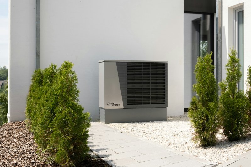 Modern Alpha Innotec air-source heat pump outdoor unit mounted on a residential wall, gravel base, evergreen shrubs flanking the cabinet