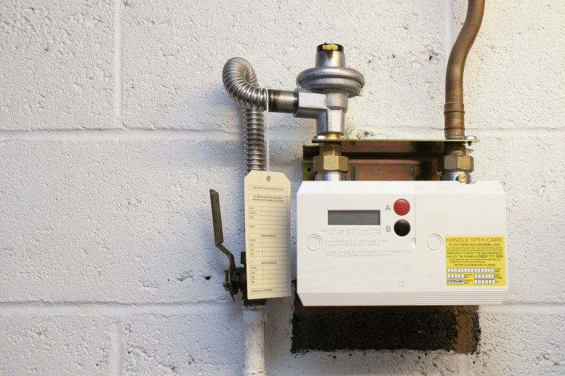 Wall-mounted residential gas meter with a yellow handle-with-care warning label, supply valve, and flexible metal connector