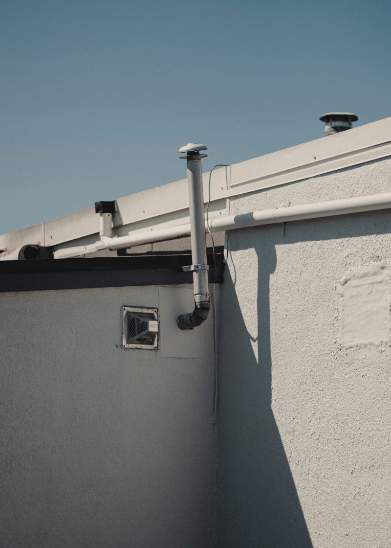 Exterior wall of a residential building with a HOT-labeled tankless-style vent termination and the PVC supply pipe routed along the stucco