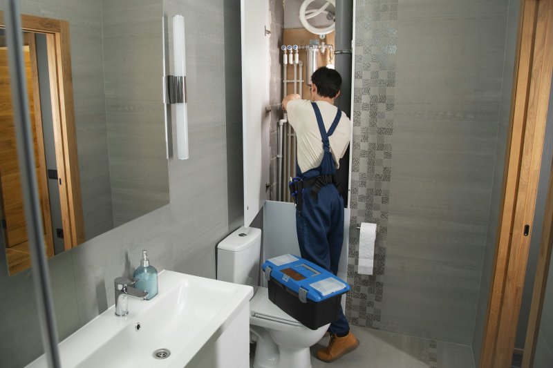 Plumber in blue overalls connecting supply pipes to a silver wall-mounted tankless water heater during a part-swap service call