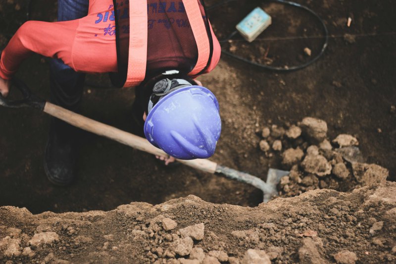 Plumber in hard hat and safety vest hand-digging a shallow spot-repair trench to expose a residential sewer line