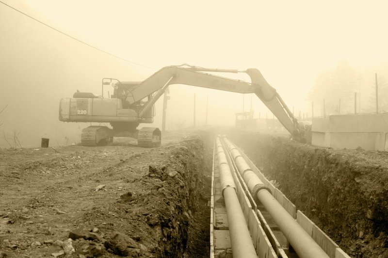 Yellow excavator digging an open trench alongside large white sewer pipes at a construction site