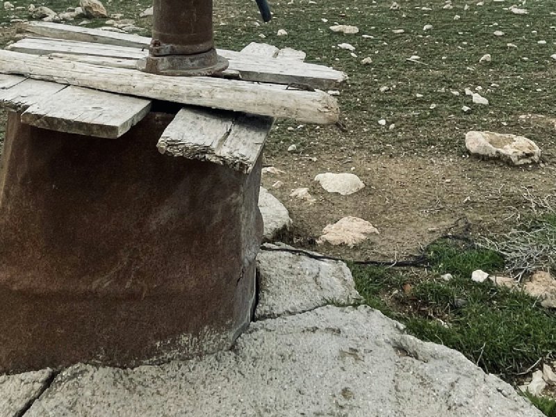 Rusty hand pump on a wooden platform at a rural well with farmhouse in the background