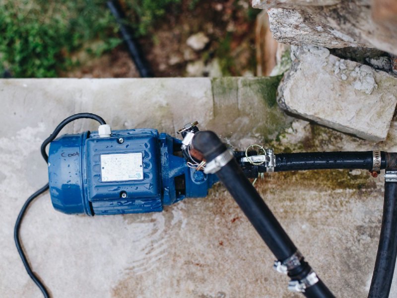 Blue residential water pump with black supply hoses and water droplets on a weathered concrete surface — the above-ground component swapped in a typical well-pump replacement