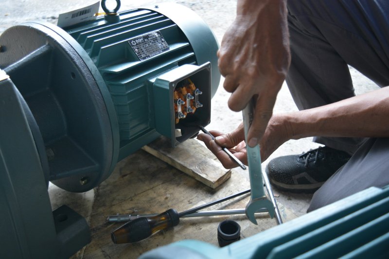 Hands repairing an industrial motor with a screwdriver and wrench, terminal box open with yellow electrical connectors