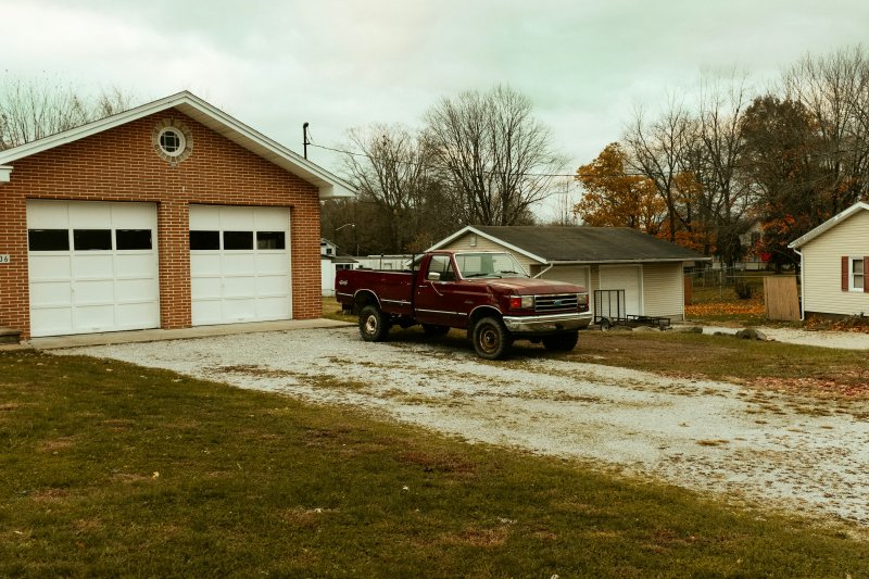 Red service pickup truck parked on a gravel driveway in front of a brick garage with two white garage doors