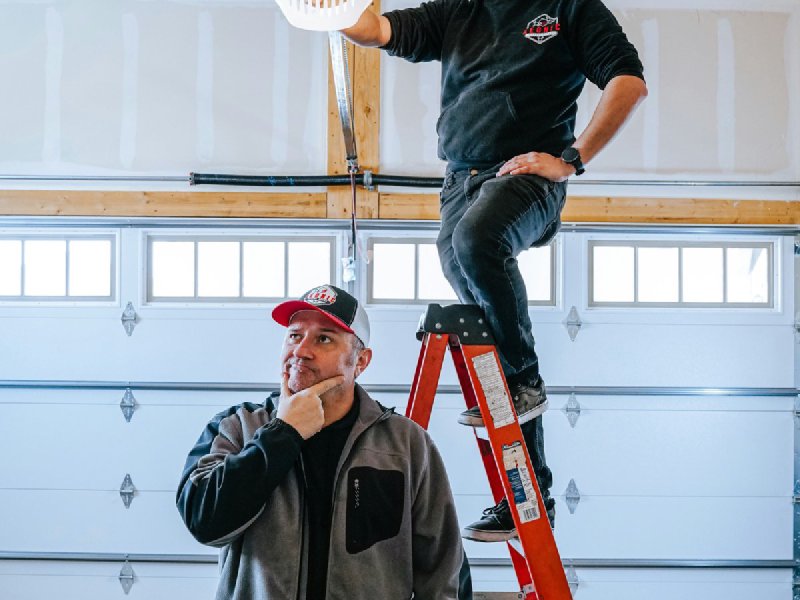 Two garage door technicians installing an opener unit on a residential ceiling, one on a ladder holding the head, the other handing up parts