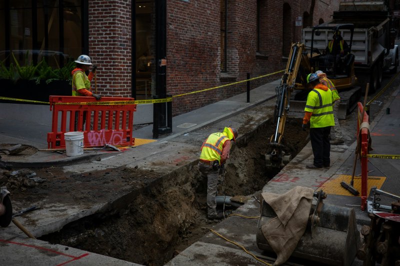 Crew in safety vests working in an open trench beside a building foundation with an excavator alongside