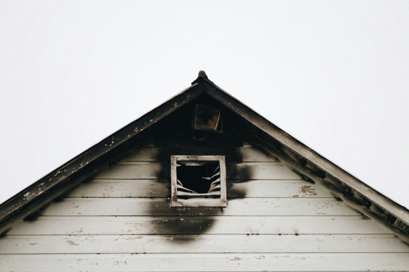 Fire-damaged white wood-frame house with blackened roof, broken window, and smoke staining around the opening
