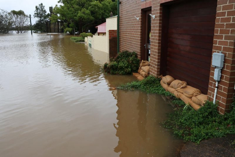 Flooded street with murky brown water reaching the foundation of a brick building, sandbags stacked along the wall to keep water out