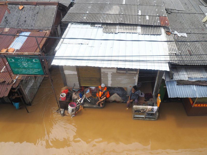 Aerial view of flooded urban street with brown floodwater submerging buildings and cars while residents perch on raised platforms