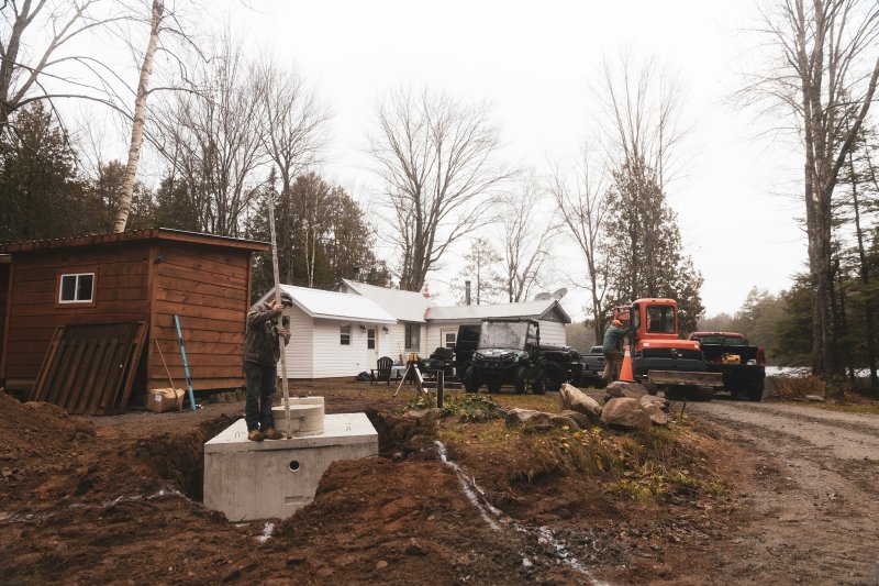 Construction worker standing on a freshly poured concrete foundation with a measuring rod, soil piles around the perimeter