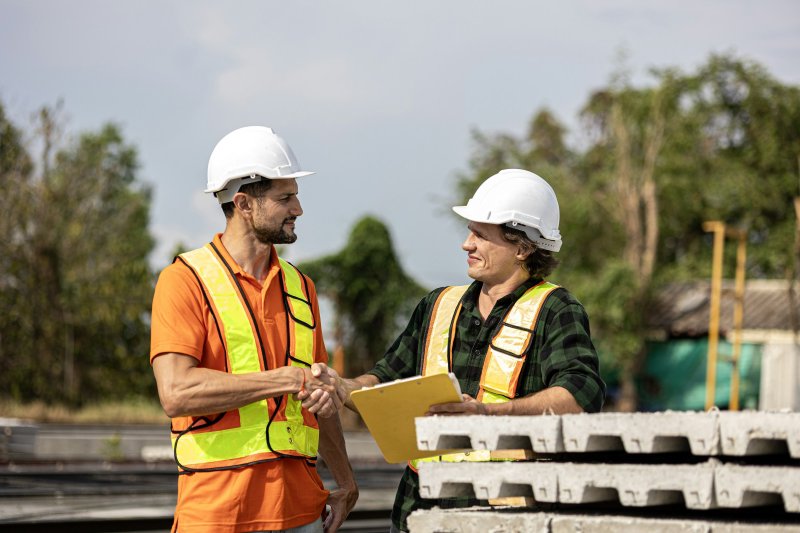Two contractors in hard hats and high-visibility vests shake hands on a residential worksite, one holding a clipboard