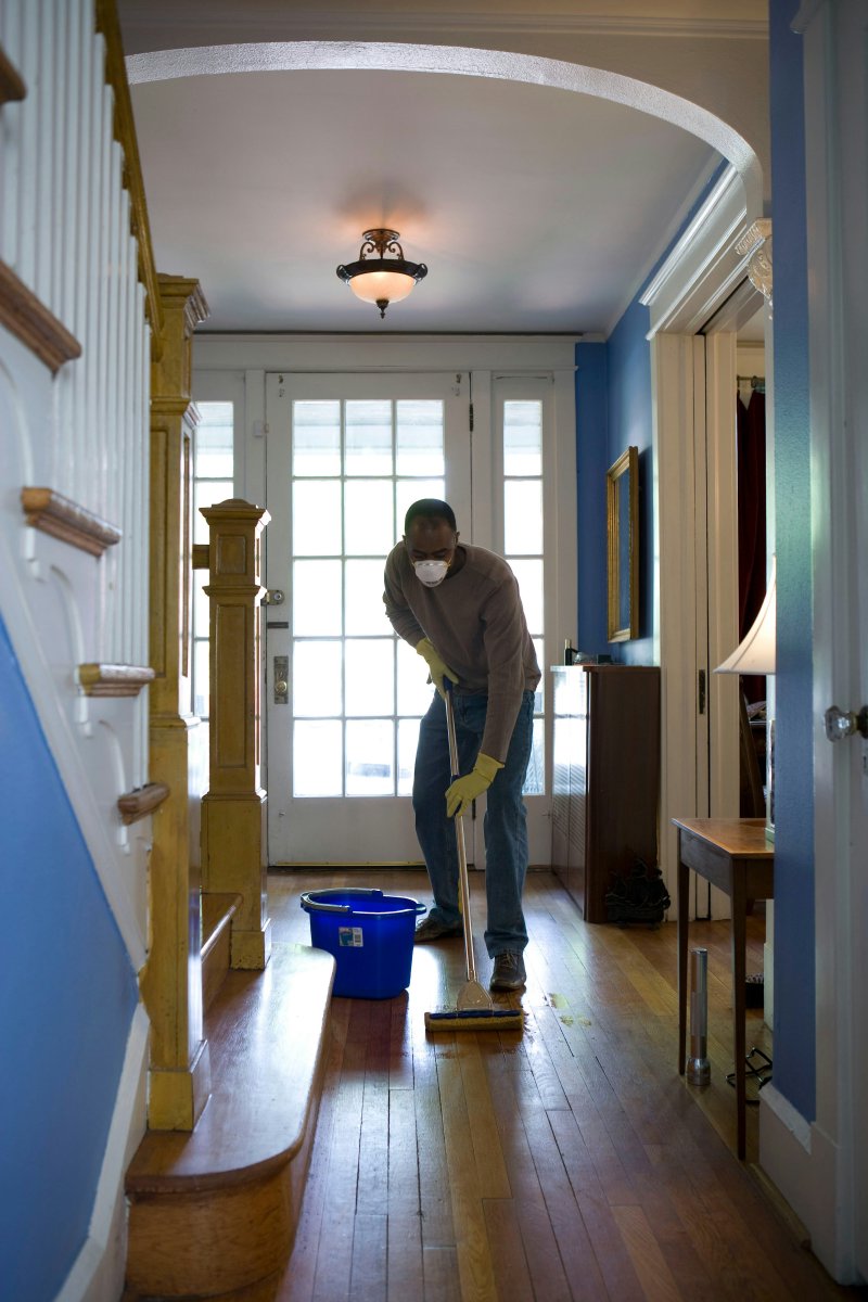 Homeowner in mask and gloves mopping a hardwood floor in a residential hallway — the DIY-scope cleanup the EPA puts under the 10-square-foot threshold
