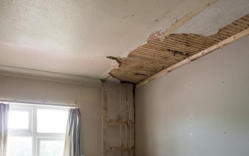 Residential room mid-restoration with damaged ceiling exposing wooden joists and insulation, furniture wrapped in protective plastic sheeting