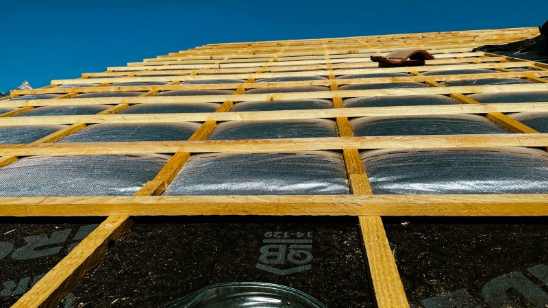 Wooden roof trusses with insulation and tar paper underlayment exposed during construction, showing the layers that sit between the deck and the shingles