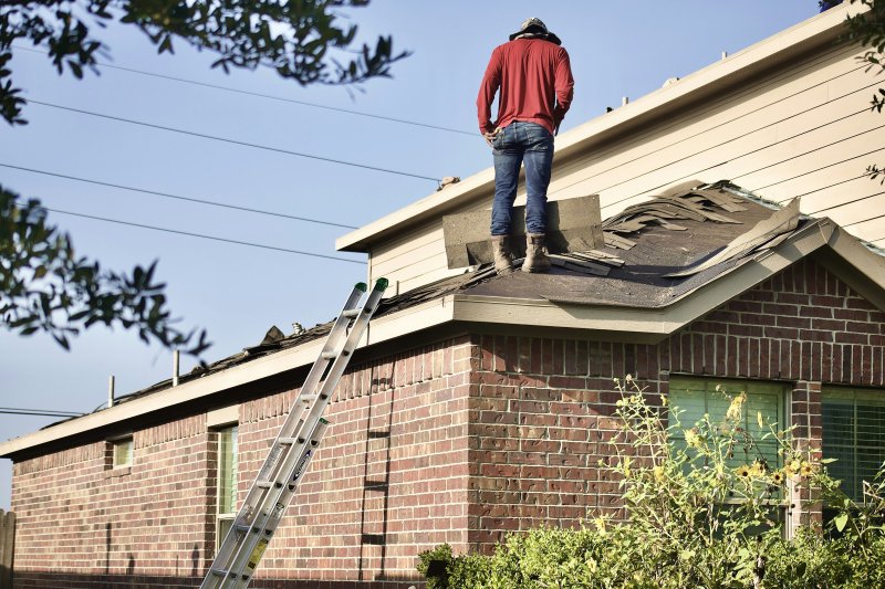Roofer in red shirt and jeans on a sloped asphalt-shingle roof, repairing damaged sections beside a brick house
