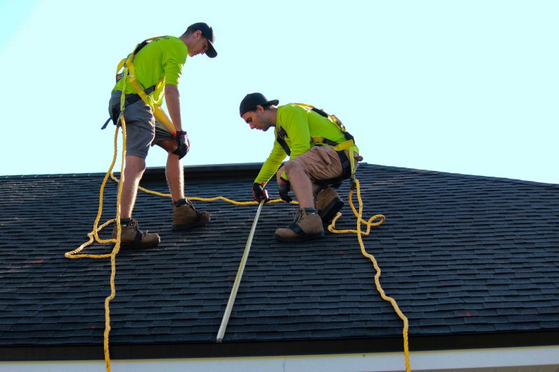 Two harnessed workers in green shirts on a dark shingled roof, one kneeling with a long measuring tape while the other holds the safety rope