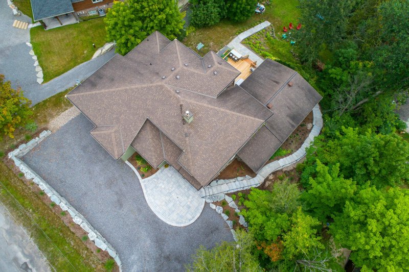 Aerial drone view of a large modern house with a complex brown shingle roof, multiple gables and dormers, surrounded by trees