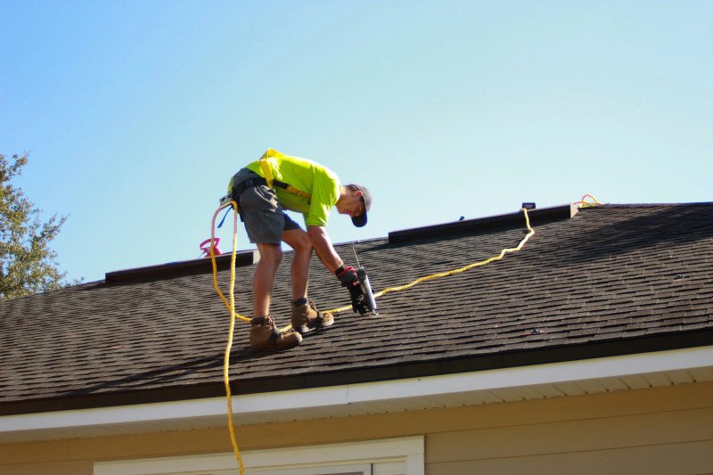 Roofer in bright yellow shirt and harness with a yellow safety rope, kneeling on a dark shingle roof while sealing with a caulking gun