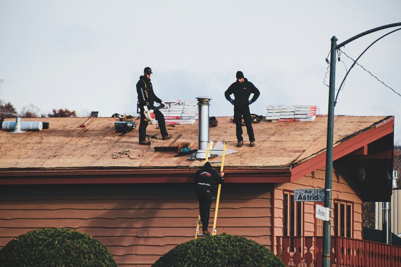 Three roofers on a residential roof installing new shingles with stacks of bundles and a yellow ladder on the eave