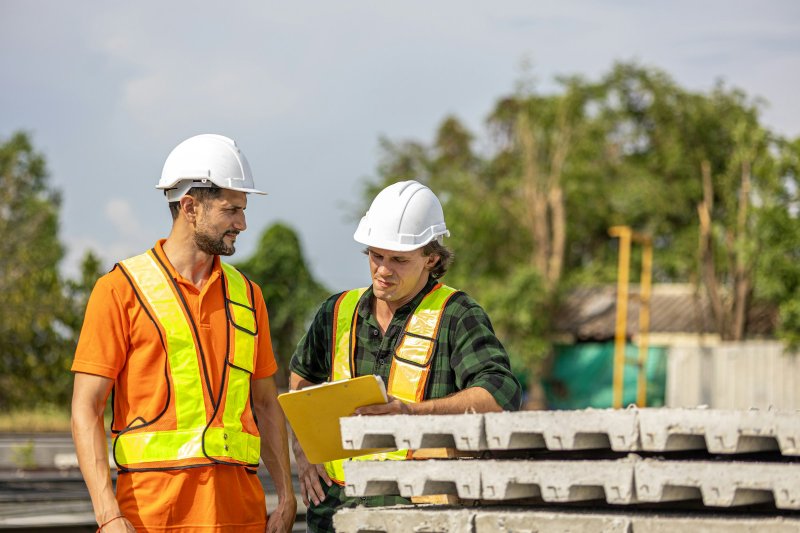 Two construction workers in white hard hats and yellow high-visibility vests reviewing project plans on a job site