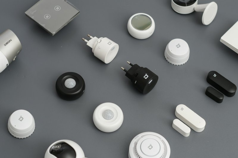 Flat-lay of smart home security devices including motion sensors, smart plugs, door and window sensors, and cameras on a gray surface